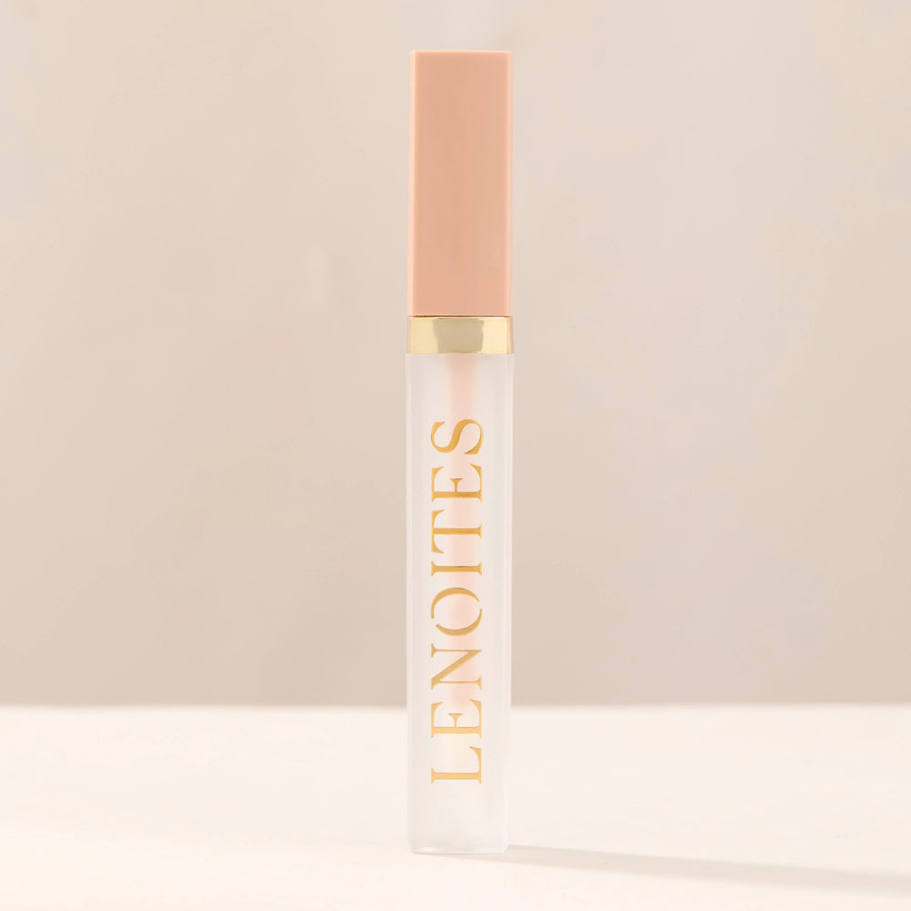 Tinted Lip Oil in Clear by Lenoites, featuring a sleek tube with a pink cap and clear body, displayed against a soft beige background.